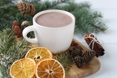 Photo of Yummy cocoa in cup, dried orange slices, cinnamon sticks and fir tree branches on white wooden table, closeup