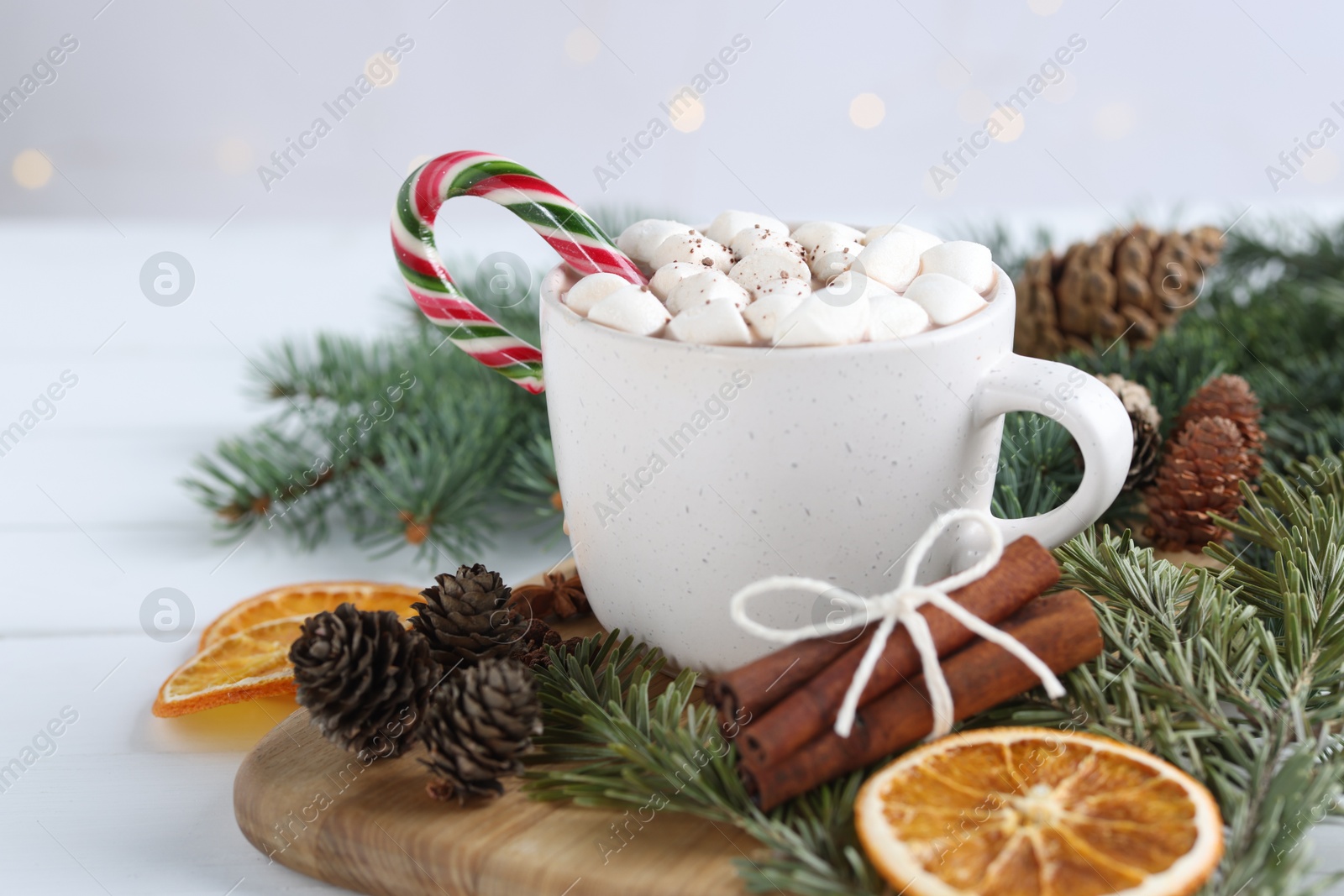 Photo of Yummy cocoa with marshmallows and candy cane in cup, dried orange slices, cinnamon sticks and fir tree branches on white table, closeup