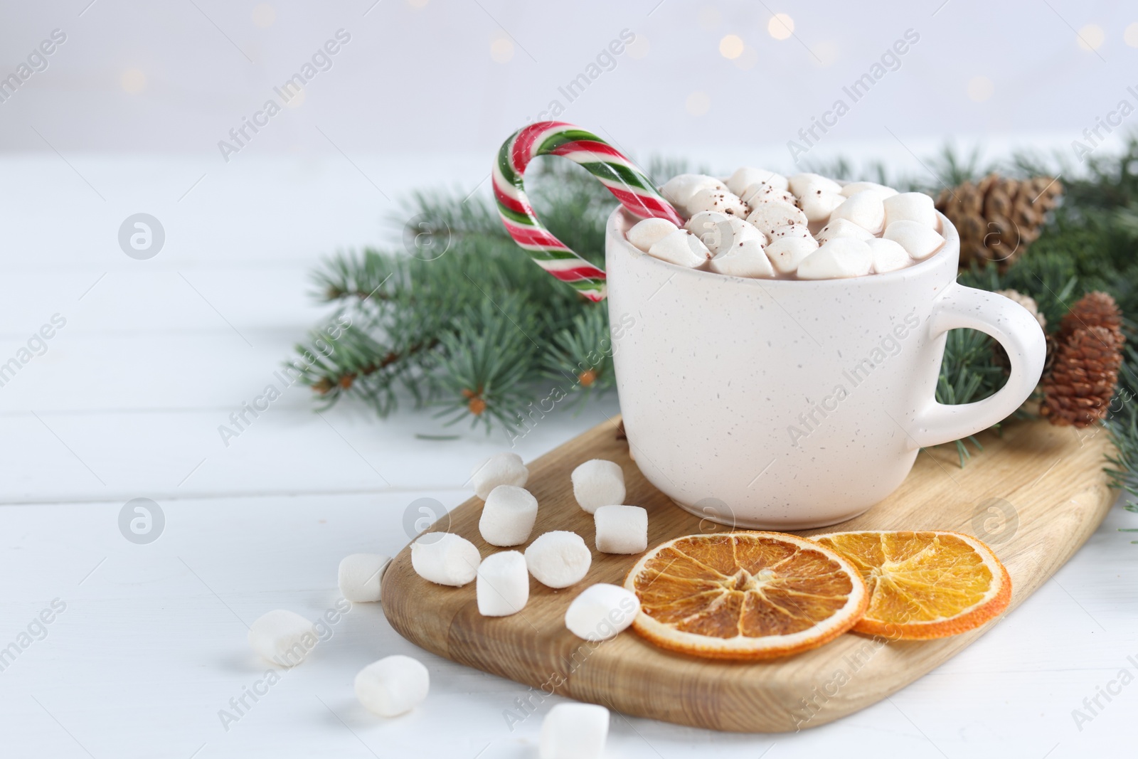 Photo of Yummy cocoa with marshmallows and candy cane in cup, dried orange slices and fir tree branches on white wooden table. Space for text