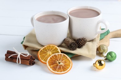 Photo of Yummy cocoa in cups, dried orange slices, cinnamon sticks and Christmas balls on white wooden table