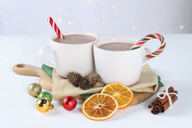 Yummy cocoa and candy canes in cups, dried orange slices, cinnamon sticks and Christmas balls on white wooden table