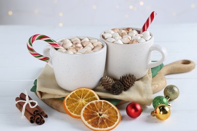 Yummy cocoa with marshmallows and candy canes in cups, dried orange slices, cinnamon sticks and Christmas balls on white wooden table