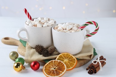 Photo of Yummy cocoa with marshmallows and candy canes in cups, dried orange slices, cinnamon sticks and Christmas balls on white wooden table