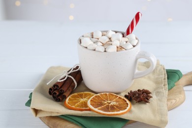 Photo of Yummy cocoa with marshmallows and candy cane in cup, dried orange slices and cinnamon sticks on white wooden table