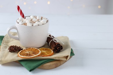 Photo of Yummy cocoa with marshmallows and candy cane in cup, dried orange slices and cinnamon sticks on white wooden table. Space for text