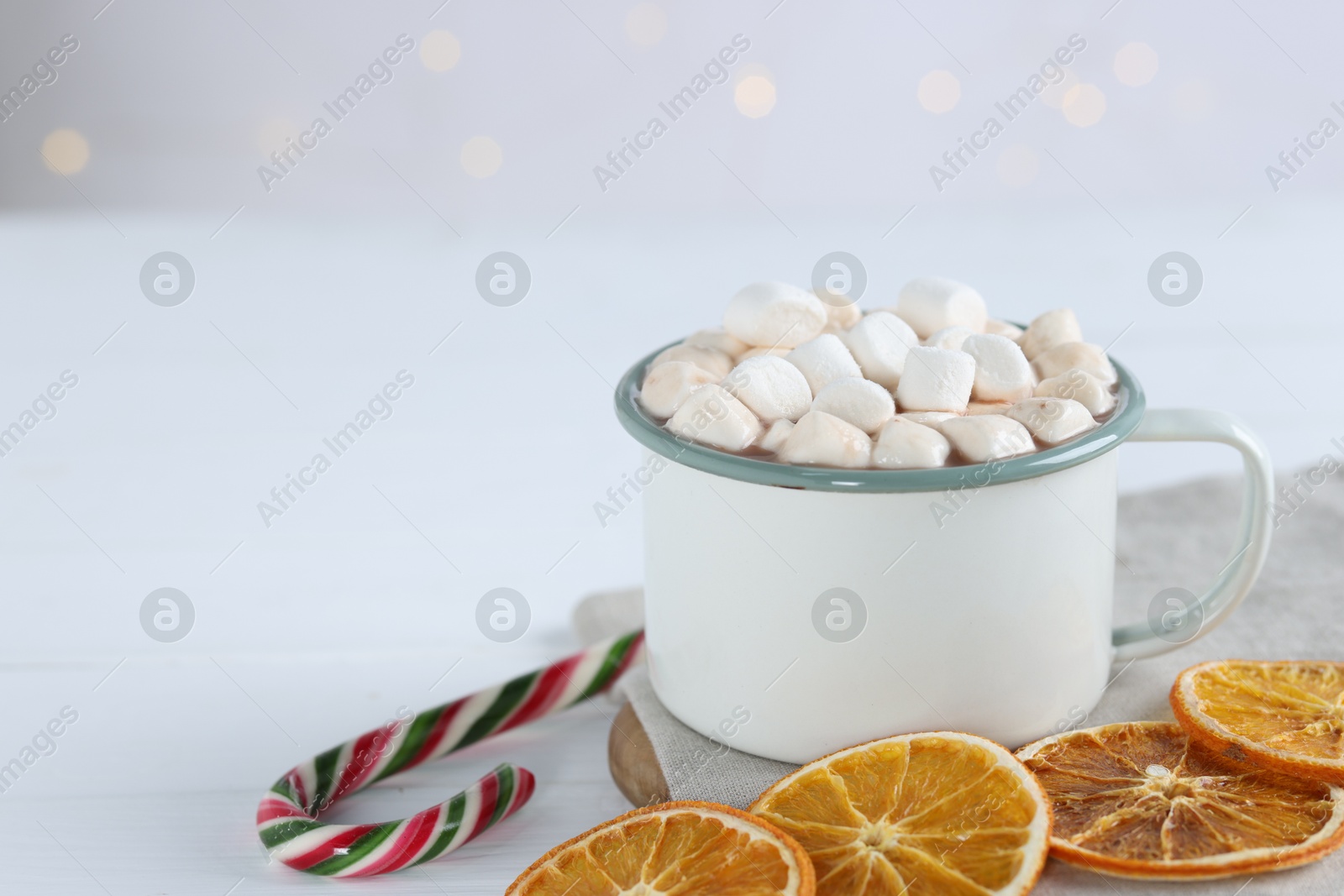 Photo of Yummy cocoa with marshmallows in cup, candy cane and dried orange slices on white wooden table. Space for text