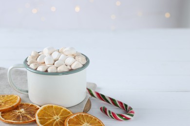 Photo of Yummy cocoa with marshmallows in cup, candy cane and dried orange slices on white wooden table. Space for text