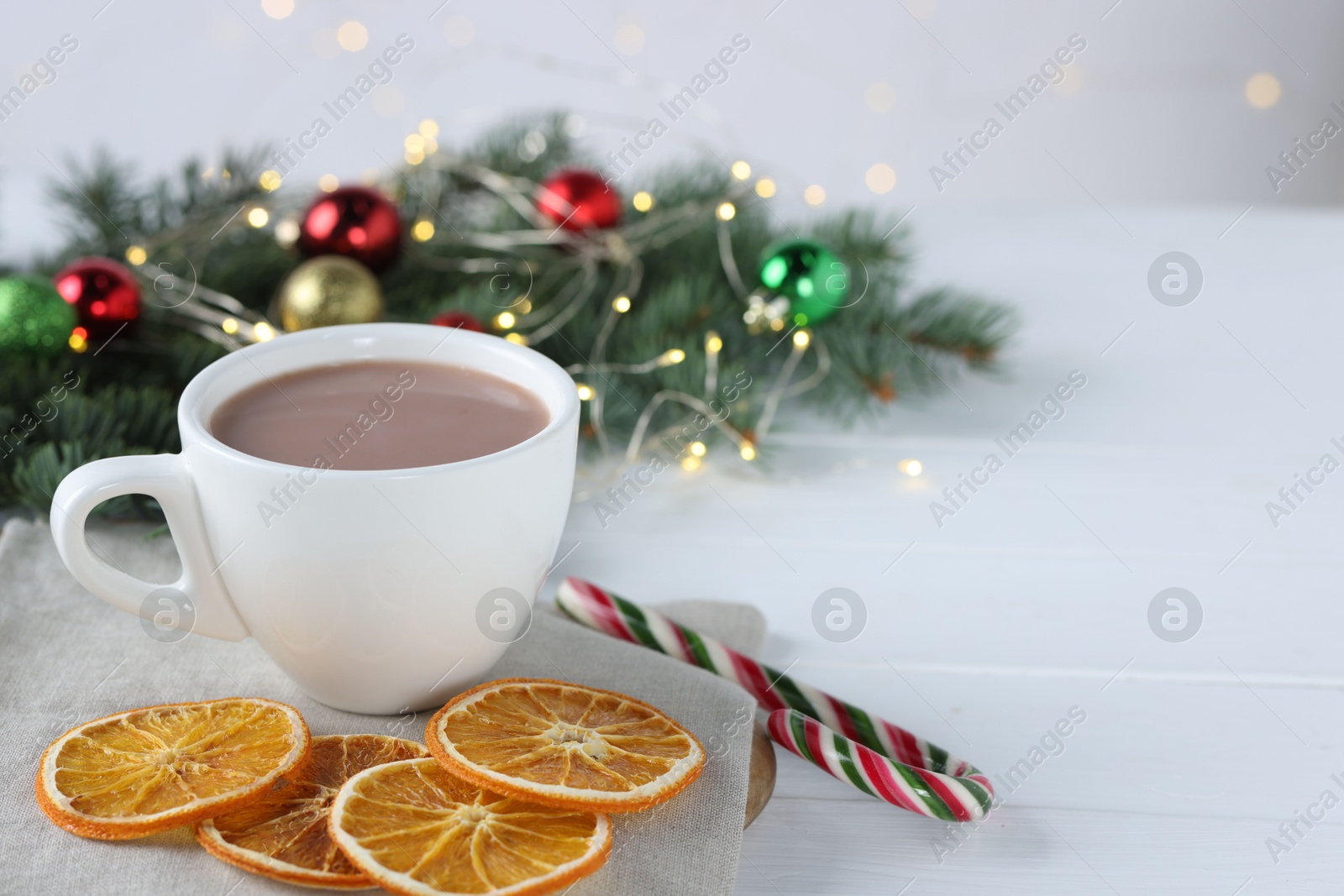 Photo of Yummy cocoa in cup, candy cane, dried orange slices and Christmas wreath on white wooden table. Space for text