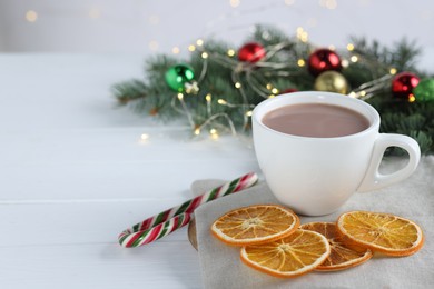 Photo of Yummy cocoa in cup, candy cane, dried orange slices and Christmas wreath on white wooden table. Space for text