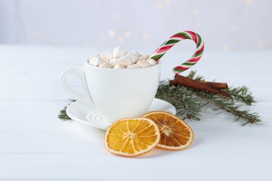 Photo of Yummy cocoa with marshmallows and candy cane in cup, dried orange slices and fir tree branches on white wooden table