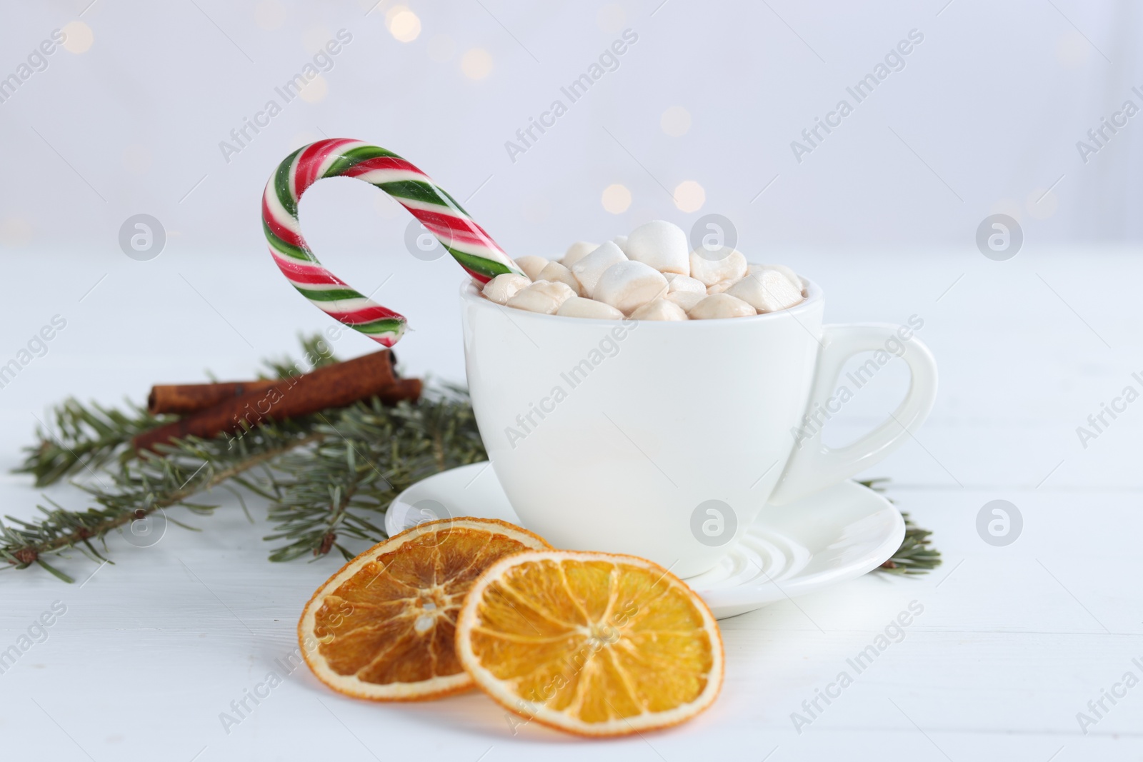 Photo of Yummy cocoa with marshmallows and candy cane in cup, dried orange slices and fir tree branches on white wooden table