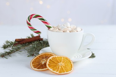 Photo of Yummy cocoa with marshmallows and candy cane in cup, dried orange slices and fir tree branches on white wooden table