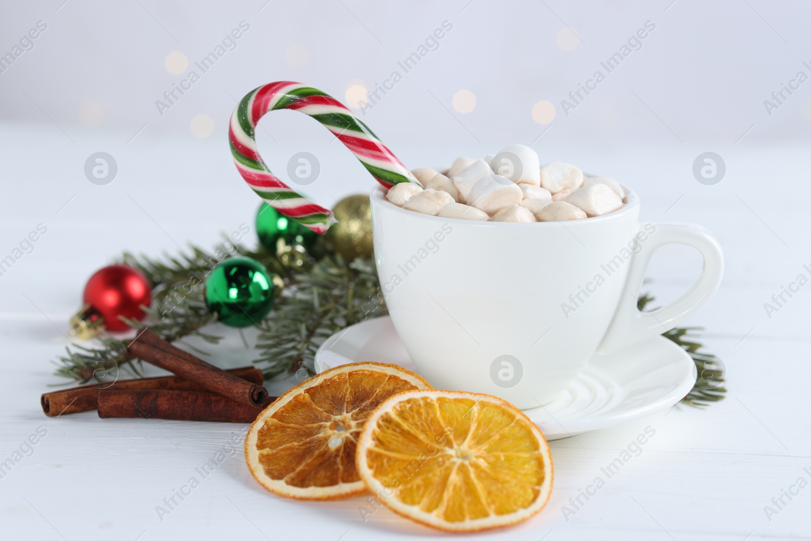 Photo of Yummy cocoa with marshmallows and candy cane in cup, dried orange slices, cinnamon sticks and fir tree branches with Christmas balls on white wooden table