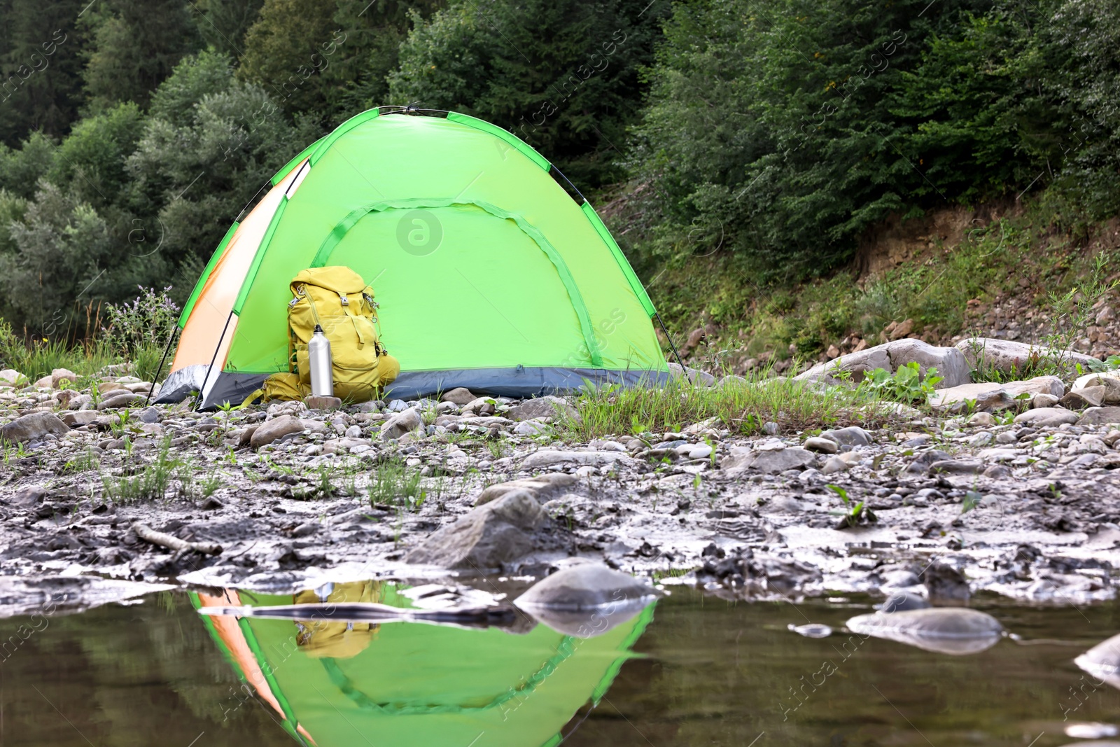 Photo of Camping tent, backpack and thermo bottle on stones near river in mountains