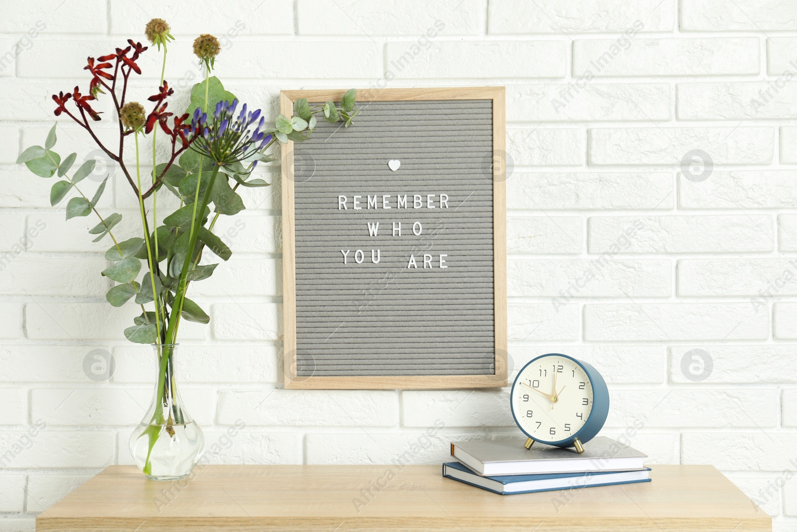 Photo of Letter board with phrase Remember Who You Are, books, alarm clock and bouquet in vase on wooden table indoors