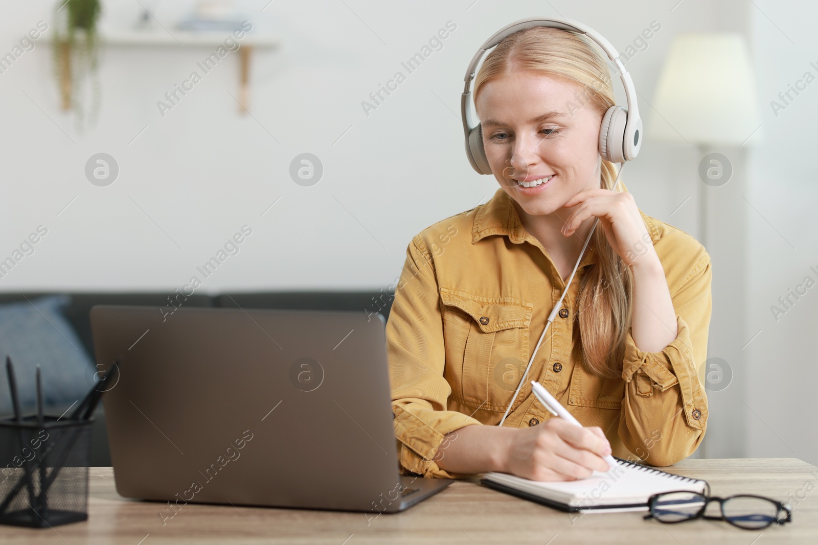 Photo of Interpreter in headphones taking notes while working with laptop at table indoors