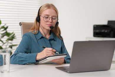 Interpreter in headset taking notes while working with laptop at table indoors