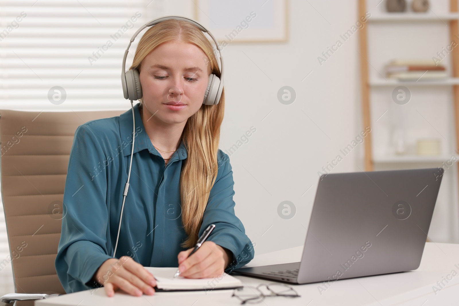 Photo of Interpreter in headphones taking notes while working with laptop at table indoors