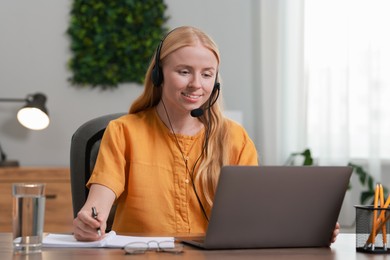 Interpreter in headset taking notes while working with laptop at table indoors