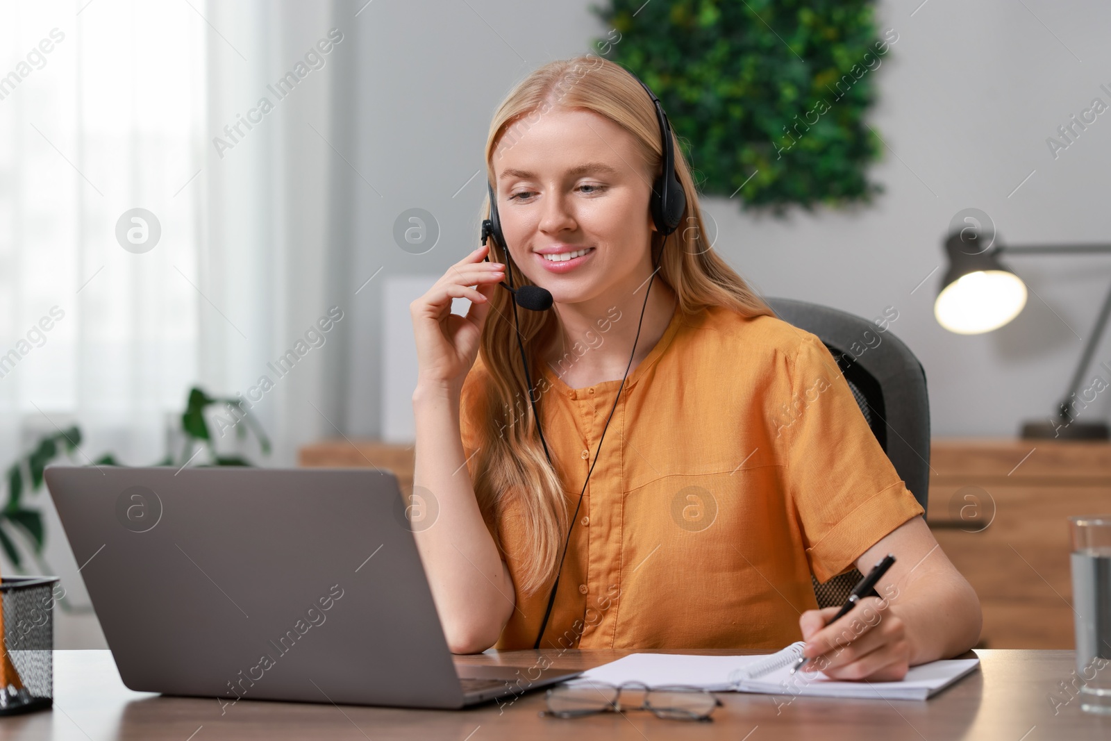 Photo of Interpreter in headphones taking notes while working with laptop at table indoors