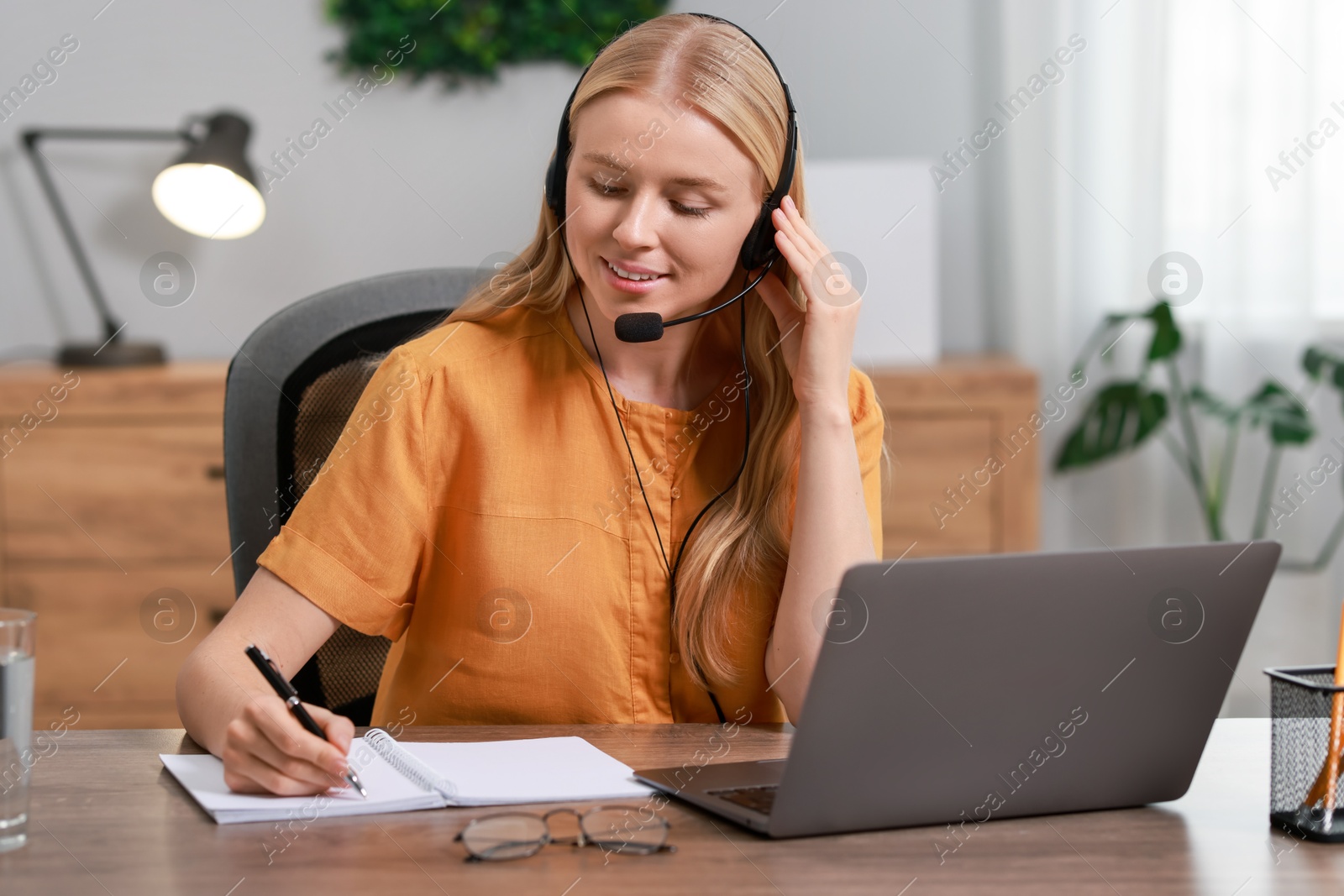 Photo of Interpreter in headphones taking notes while working with laptop at table indoors