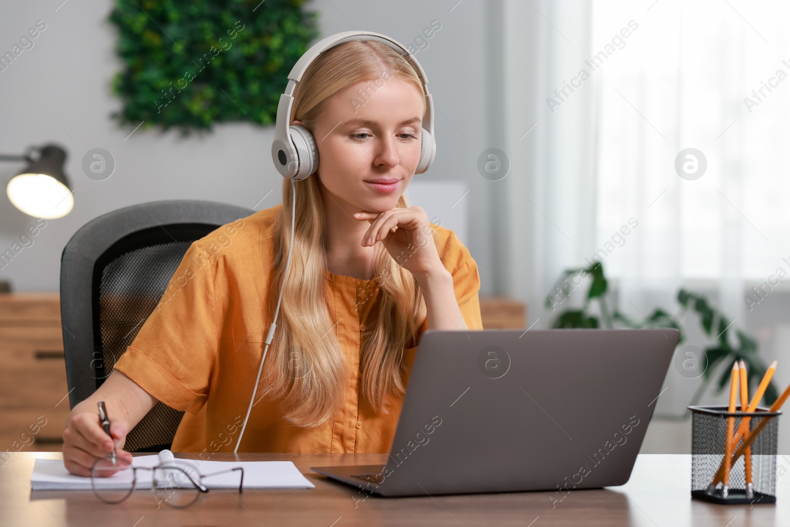 Photo of Interpreter in headphones taking notes while working with laptop at table indoors