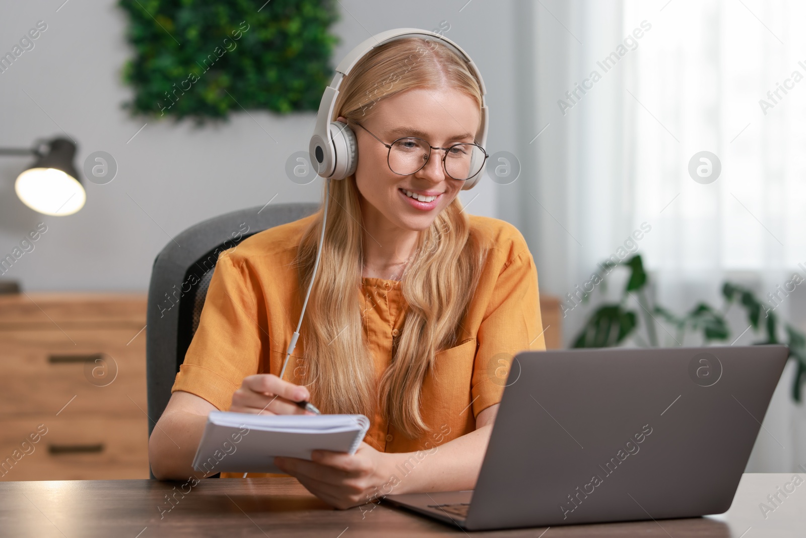 Photo of Interpreter in headphones taking notes while having video chat via laptop at table indoors