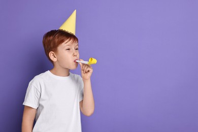 Little boy in party hat with blower on purple background, space for text