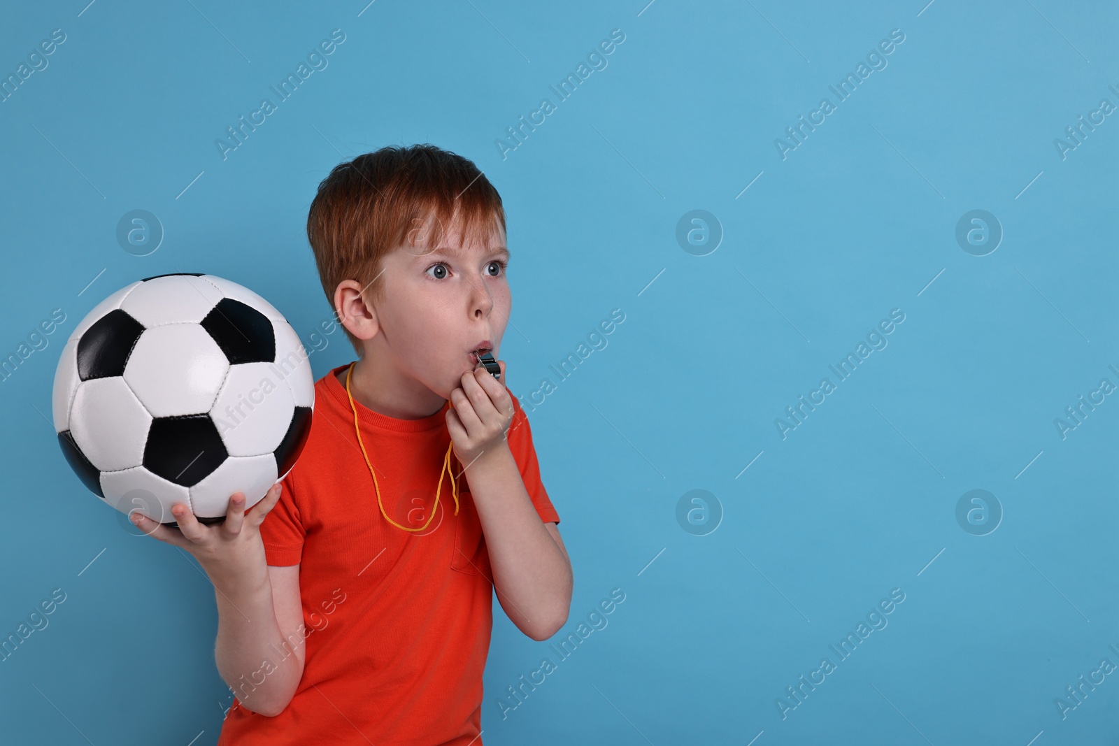 Photo of Little boy with soccer ball blowing whistle on light blue background, space for text