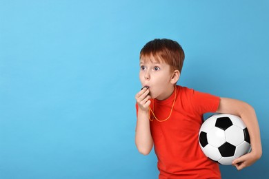 Little boy with soccer ball blowing whistle on light blue background, space for text