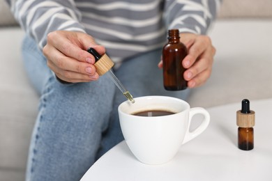 Young woman putting CBD tincture into cup with drink at white table, closeup