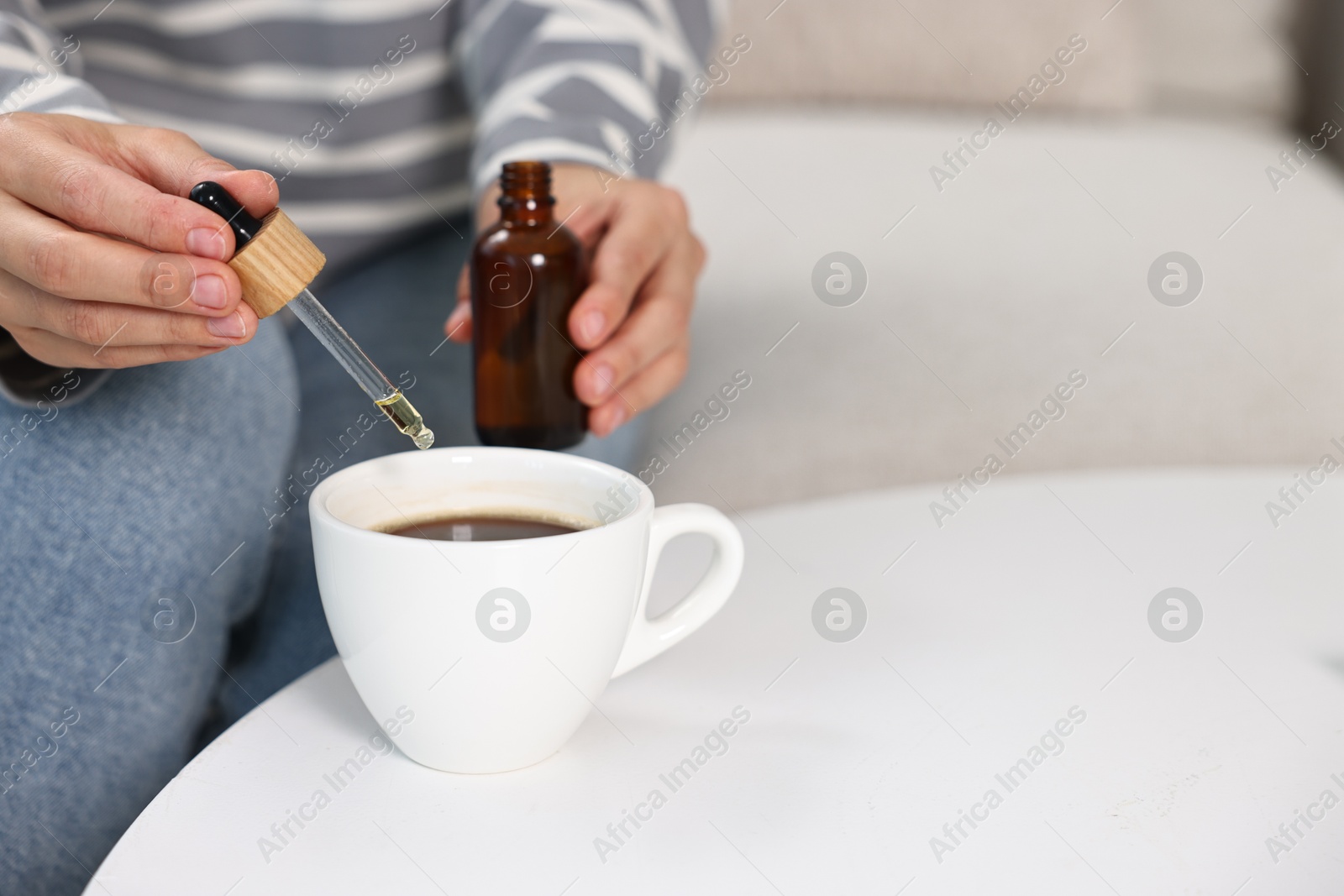 Photo of Young woman putting CBD tincture into cup with drink at white table, closeup. Space for text