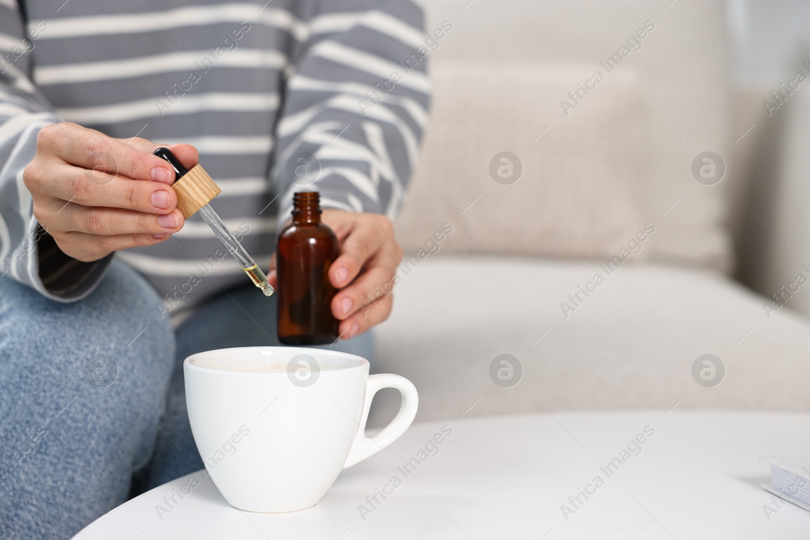 Photo of Young woman putting CBD tincture into cup with drink at white table, closeup. Space for text