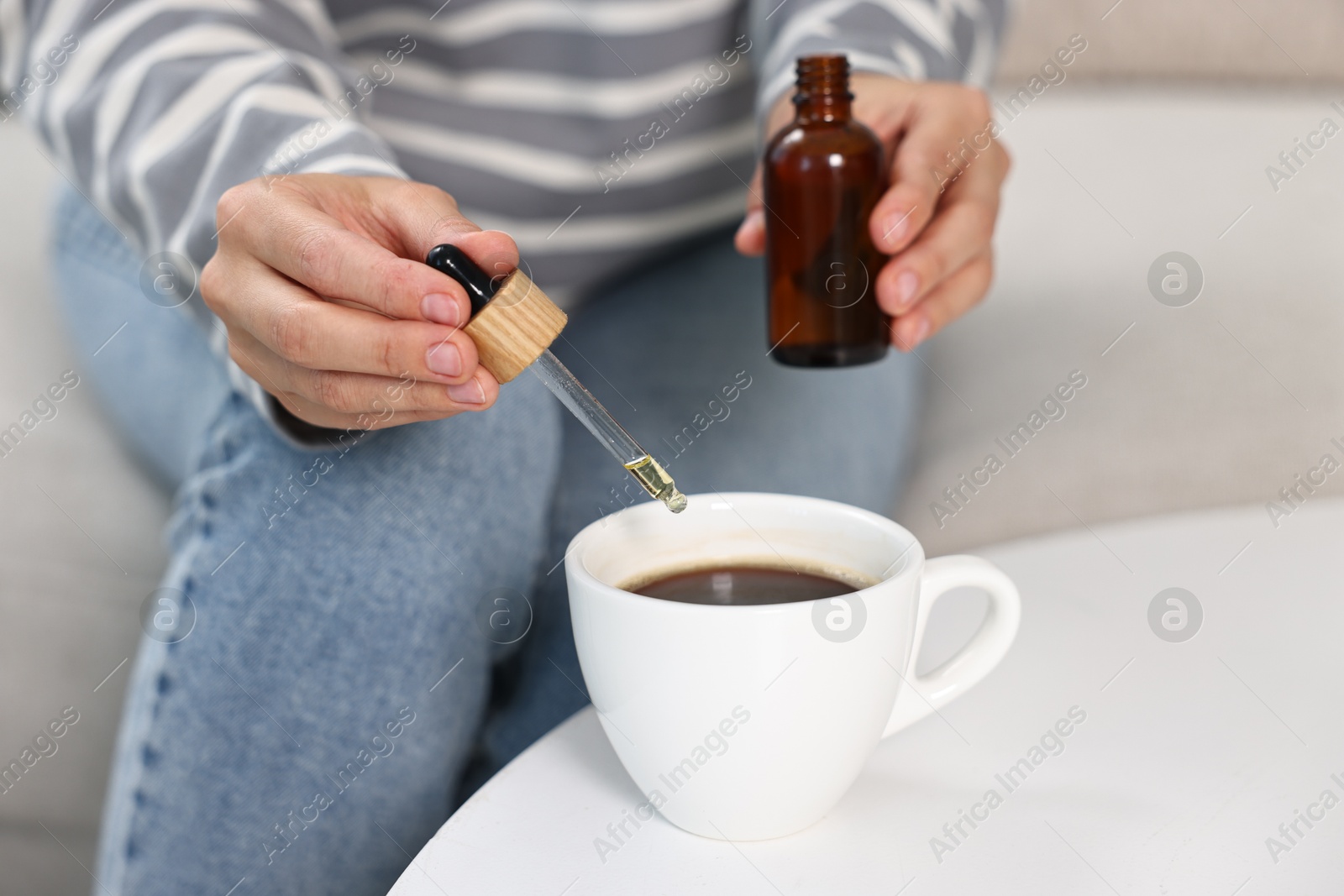 Photo of Young woman putting CBD tincture into cup with drink at white table, closeup