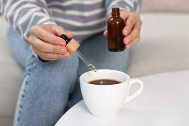 Young woman putting CBD tincture into cup with drink at white table, closeup