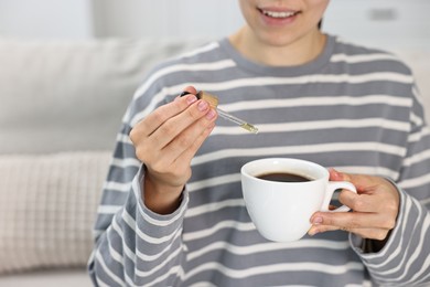 Photo of Young woman putting CBD tincture into cup with drink indoors, closeup