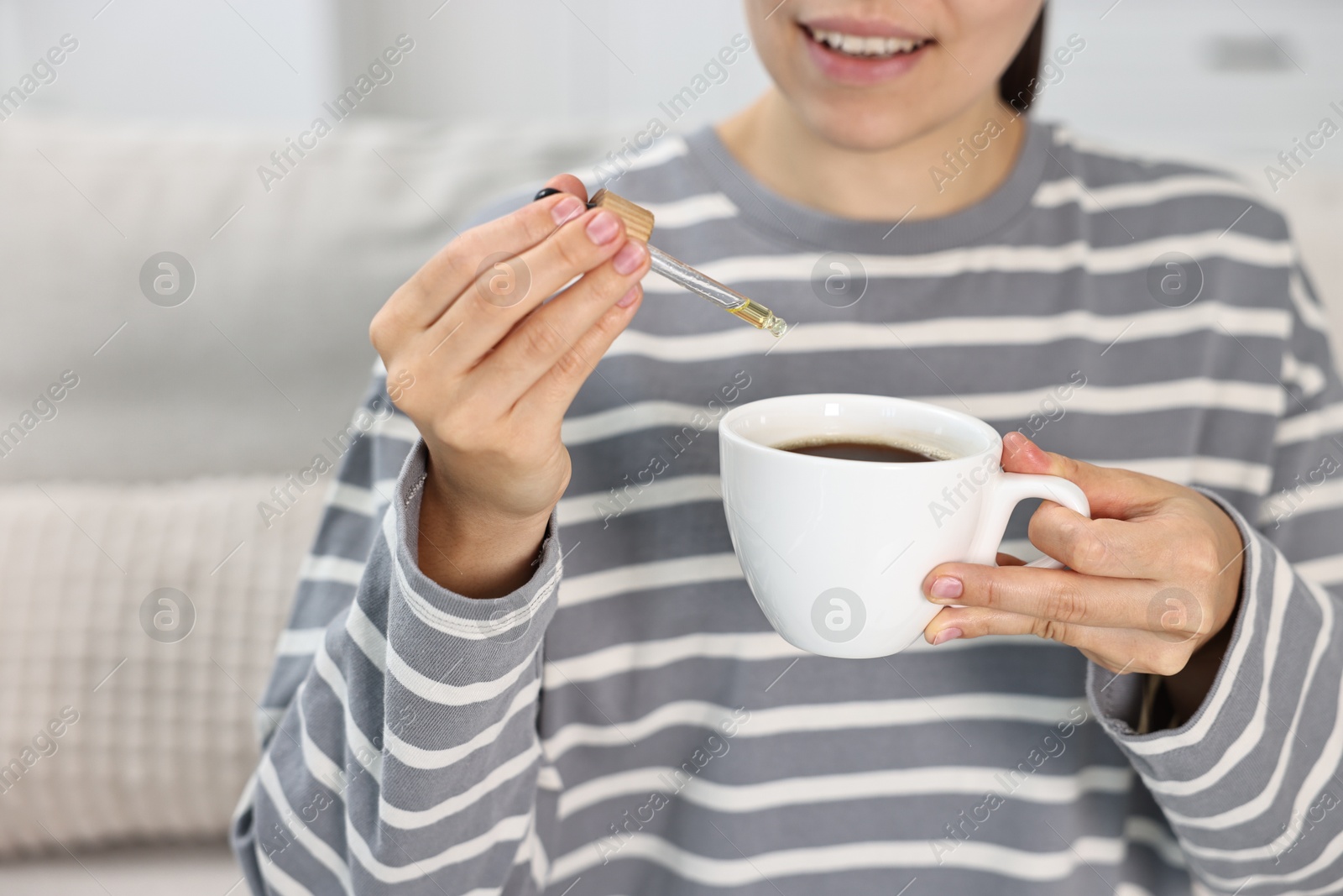 Photo of Young woman putting CBD tincture into cup with drink indoors, closeup