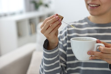 Photo of Young woman putting CBD tincture into cup with drink indoors, closeup