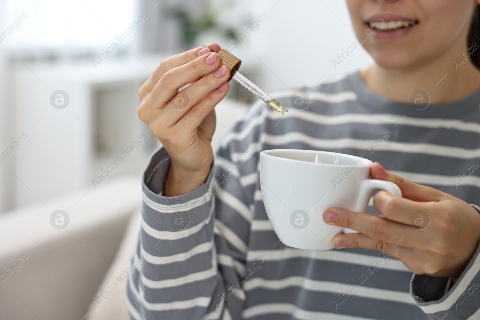 Photo of Young woman putting CBD tincture into cup with drink indoors, closeup