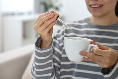 Photo of Young woman putting CBD tincture into cup with drink indoors, closeup