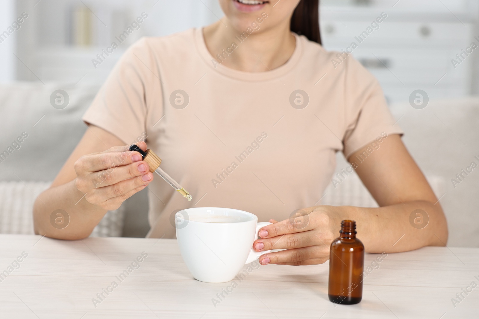 Photo of Young woman putting CBD tincture into cup with drink at white wooden table, closeup