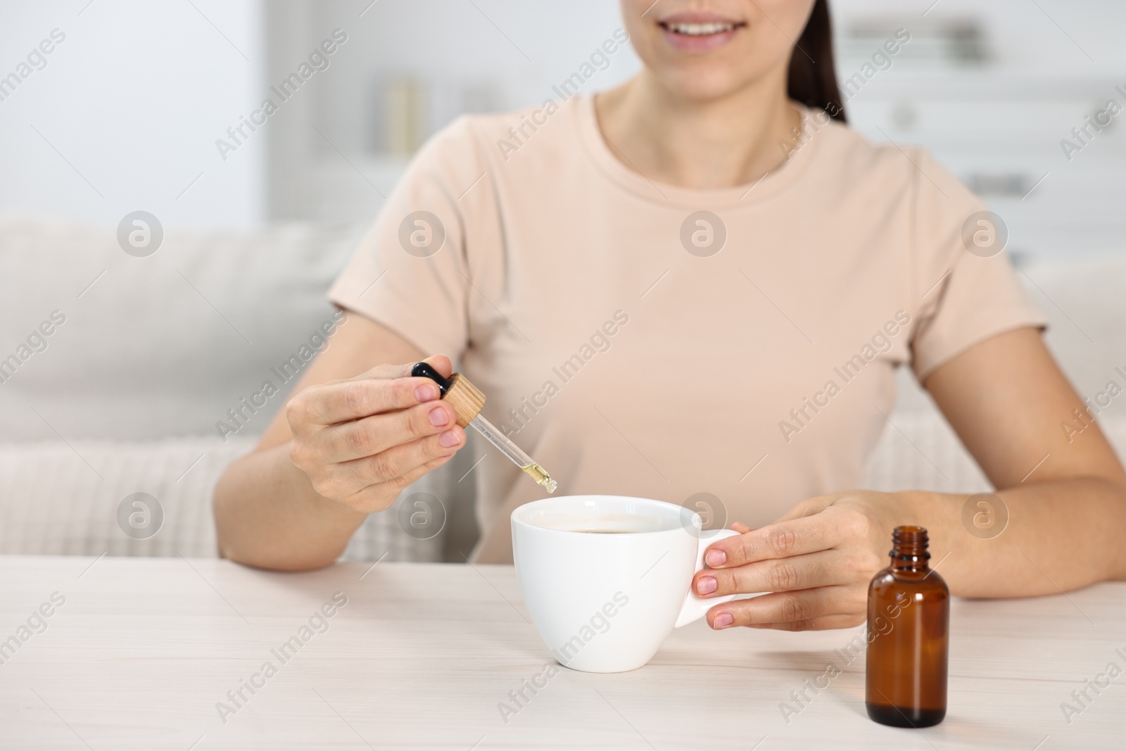 Photo of Young woman putting CBD tincture into cup with drink at white wooden table, closeup