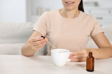 Photo of Young woman putting CBD tincture into cup with drink at white wooden table, closeup