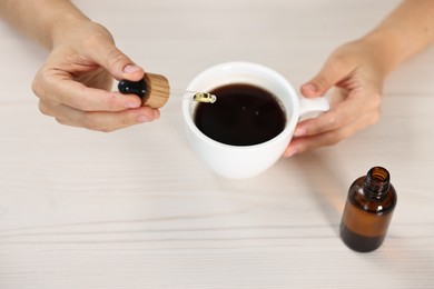 Young woman putting CBD tincture into cup with drink at white wooden table, closeup