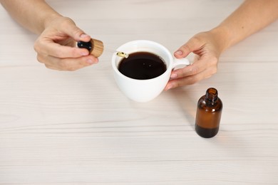 Photo of Young woman putting CBD tincture into cup with drink at white wooden table, closeup