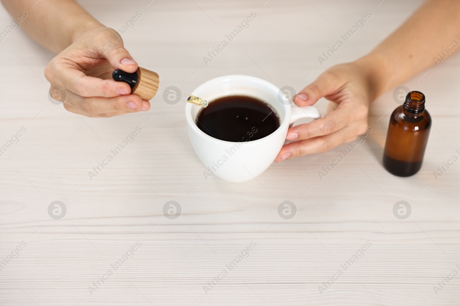 Photo of Young woman putting CBD tincture into cup with drink at white wooden table, closeup