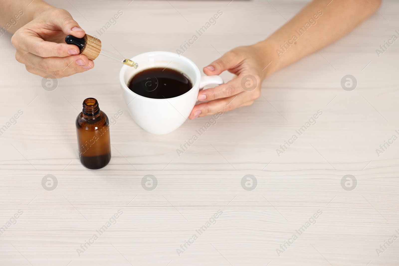 Photo of Young woman putting CBD tincture into cup with drink at white wooden table, closeup. Space for text