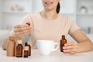 Young woman putting CBD tincture into cup with drink at white wooden table, closeup