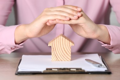 Photo of Property insurance. Real estate agent protecting wooden house figure at table indoors, closeup