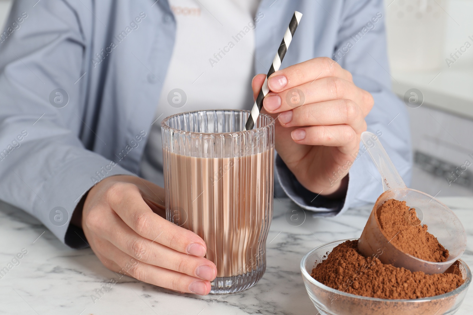 Photo of Woman with glass of protein cocktail and powder at white marble table, closeup